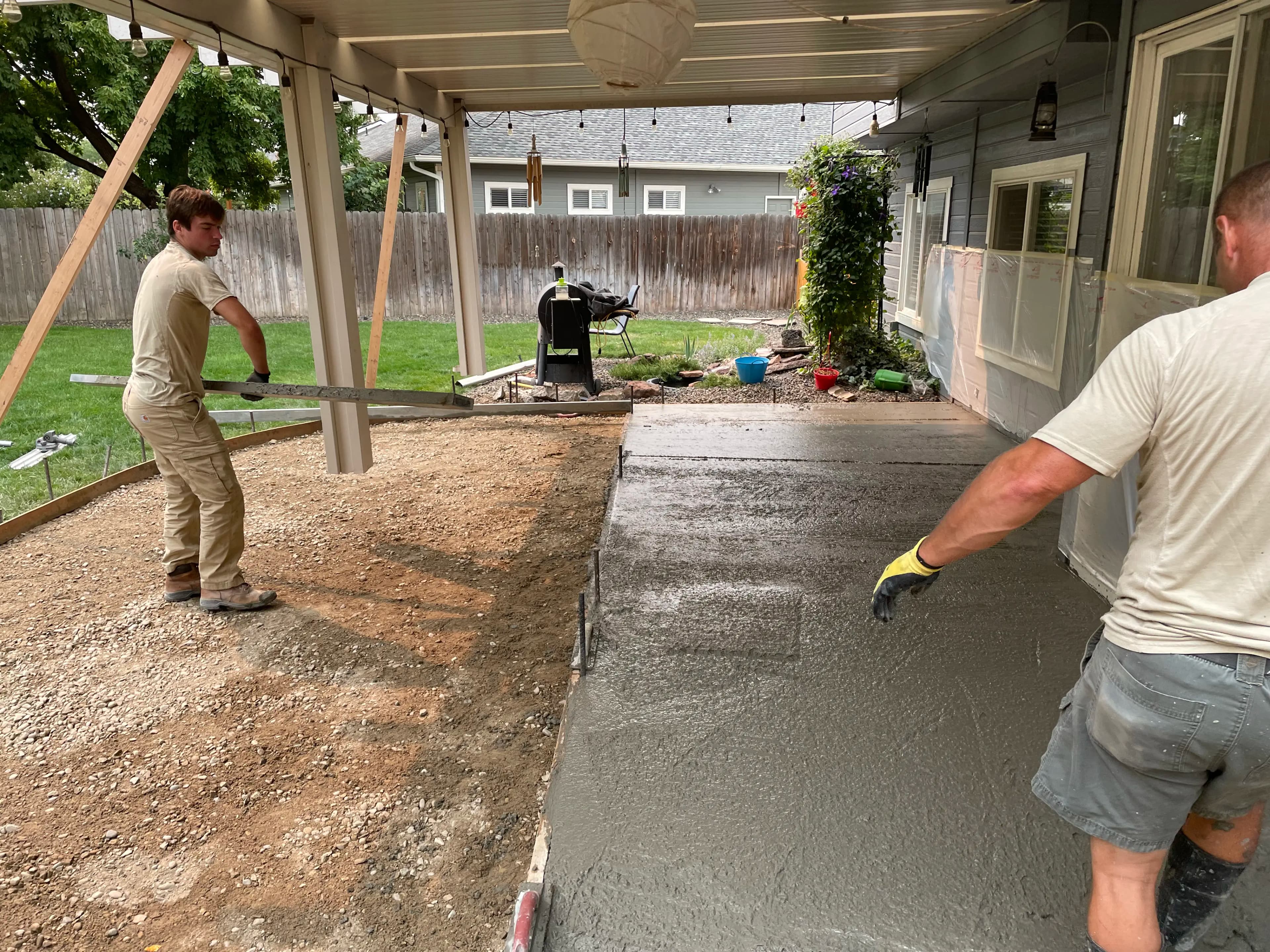 Two workers screeding wet concrete under a covered backyard patio